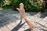 A cute child sweeping a garden pathway with a broom under sunny weather.